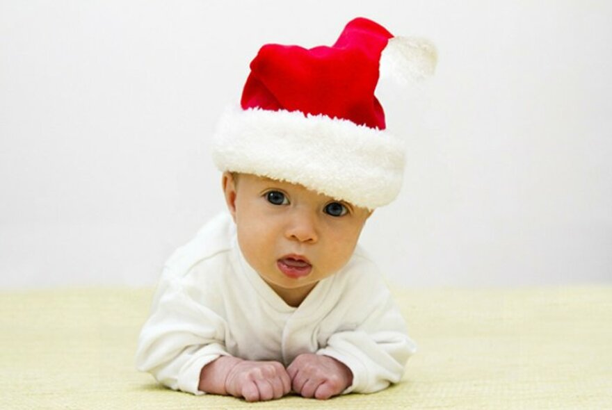 Small baby lying tummy down on a rug, upper body raised using their forearms, wearing a red and white Santa cap and a white romper.