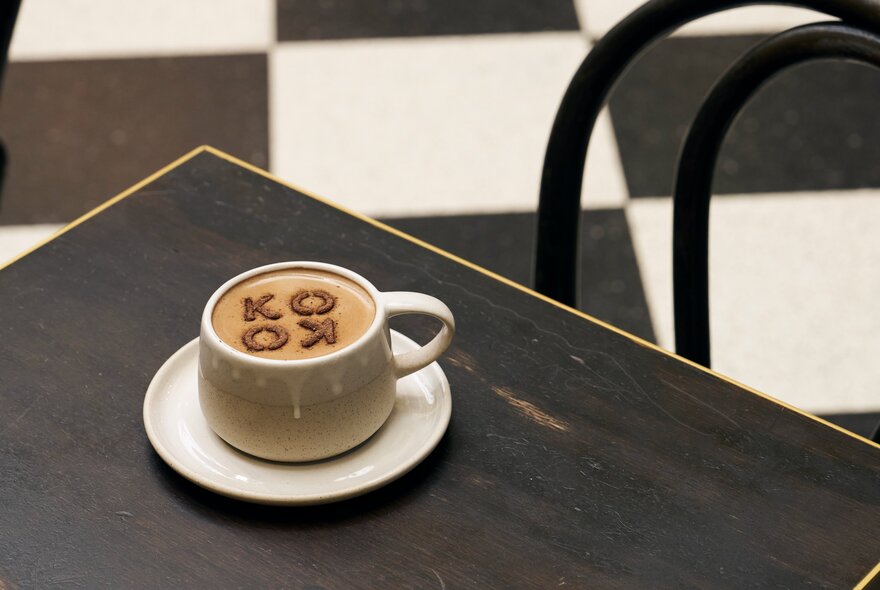 A cup of hot chocolate on a table with chocolate powder in a stencil that reads KOKO.