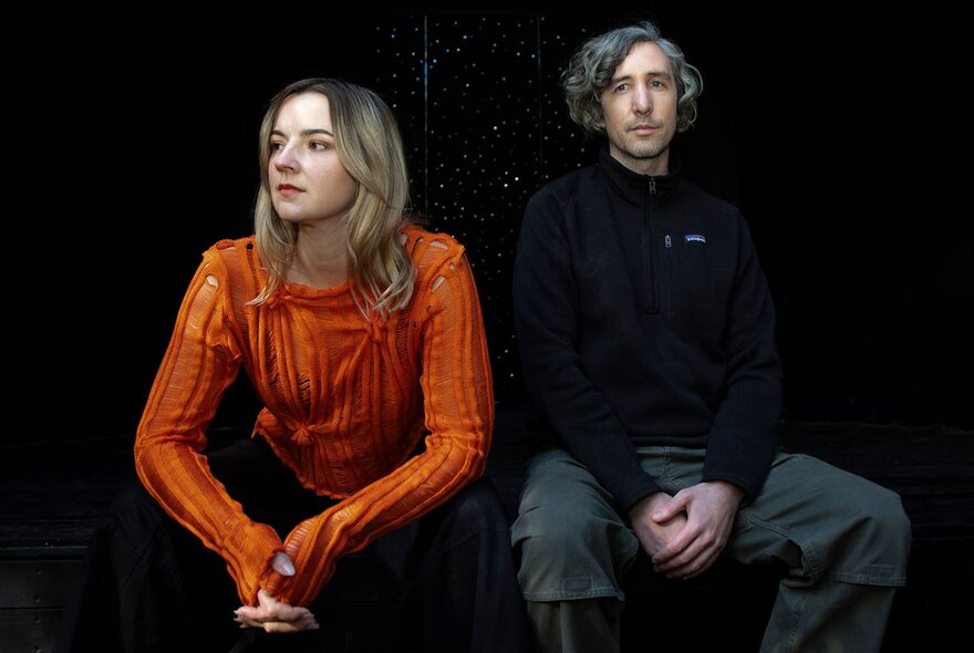 Two people posing for a photo; a woman in an orange top seated and with her hands on her knees looking off to one side and a man wearing a black jumper and grey pants leaning back against a wall, also seated.