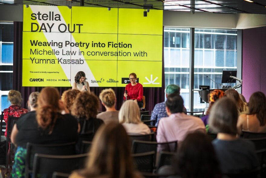 Seated audience listening to two presenters seated in front of a large screen advertising the Stella Day Out.