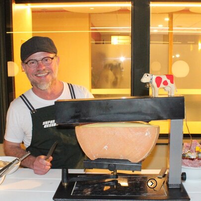 A smiling person serving raclette from a tabletop grill, in a room with a large window in the background.