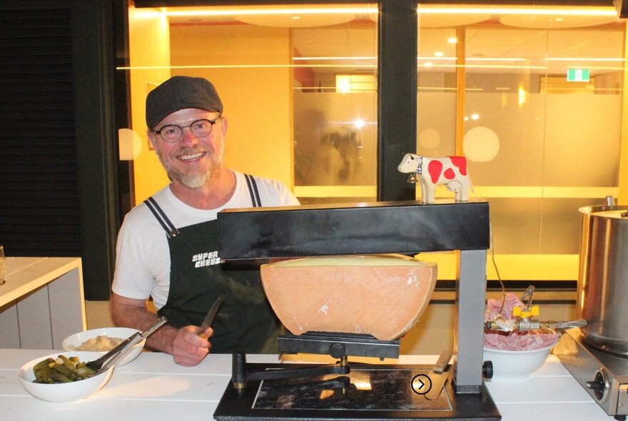 A smiling person serving raclette from a tabletop grill, in a room with a large window in the background.