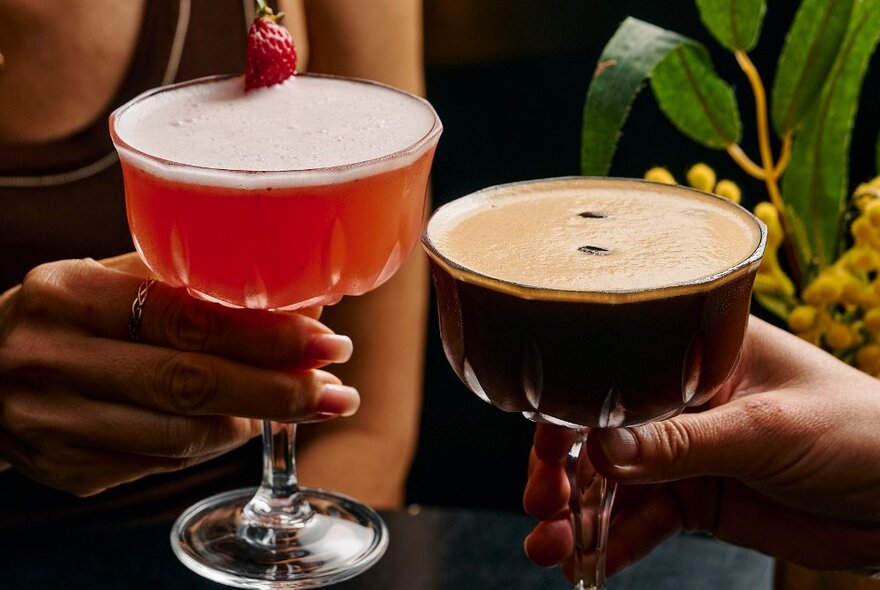 Two women's hands holding cocktails, one, an espresso martini, and the other a strawberry daquiri.