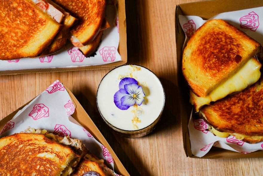 A coffee with a purple nasturtium in its centre, surrounded by various toasted sandwiches in cardboard trays.