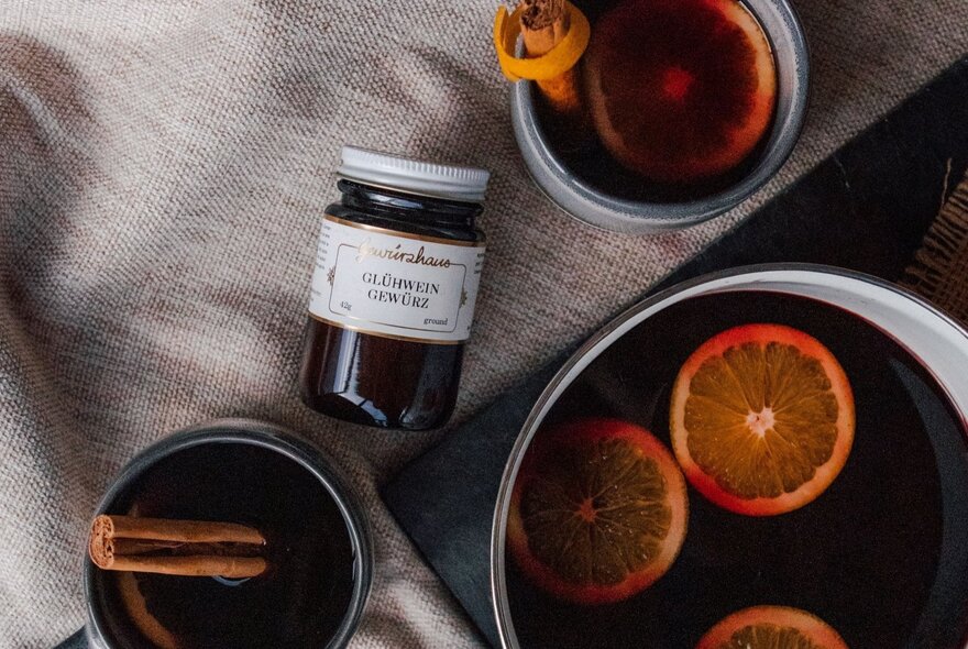 A pot of mulled wine and two mugfuls, garnished with orange slices and cinnamon, plus a jar of Glühwein spice mix.