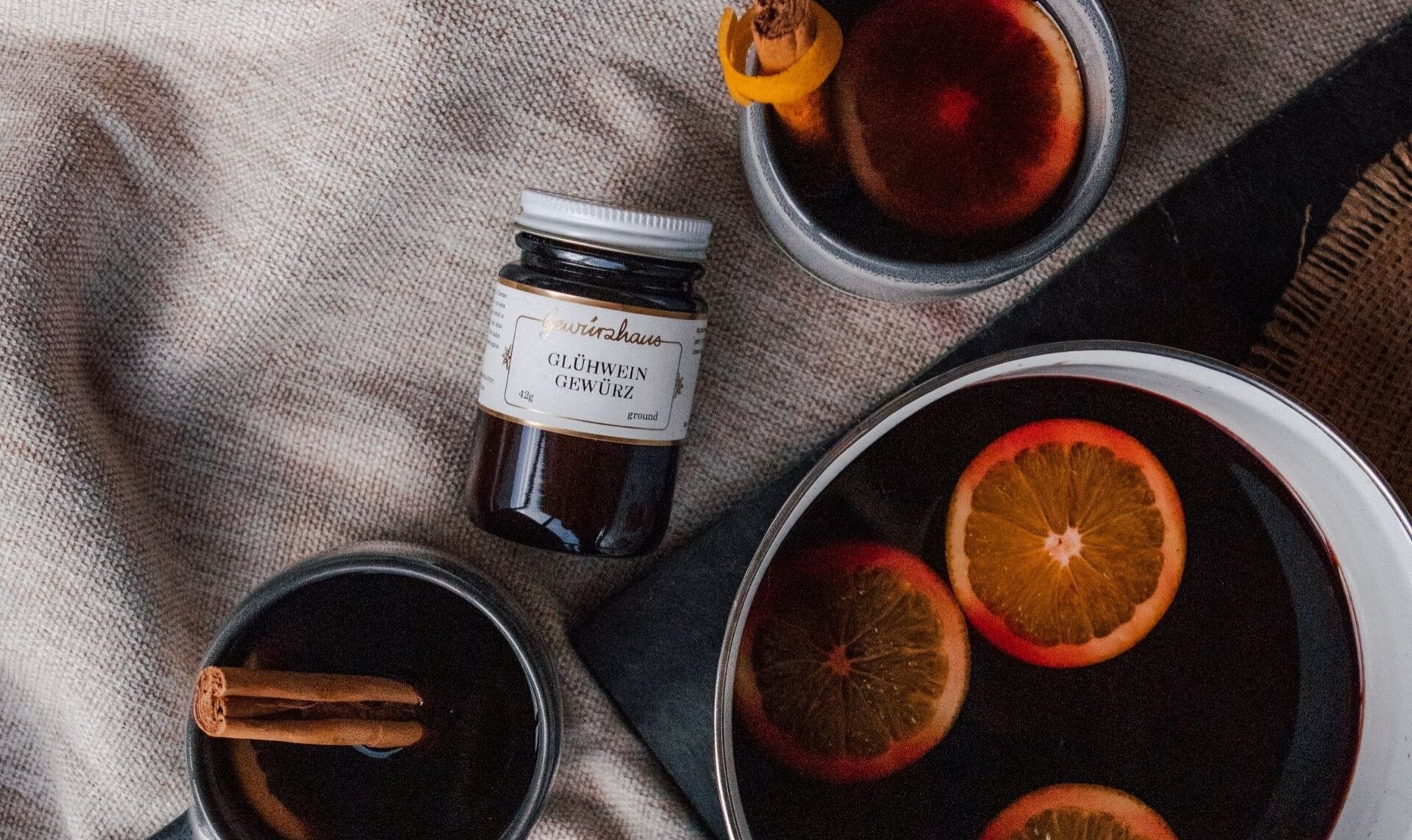 A pot of mulled wine and two mugfuls, garnished with orange slices and cinnamon, plus a jar of Glühwein spice mix.