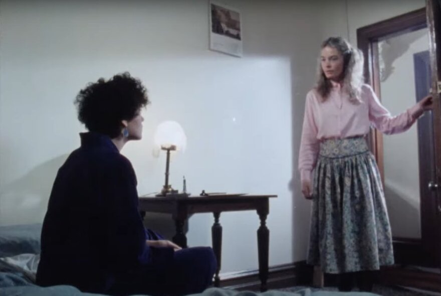 A movie still with two actors in a room, one sitting on a bed and one standing in an open doorway, both looking at each other.