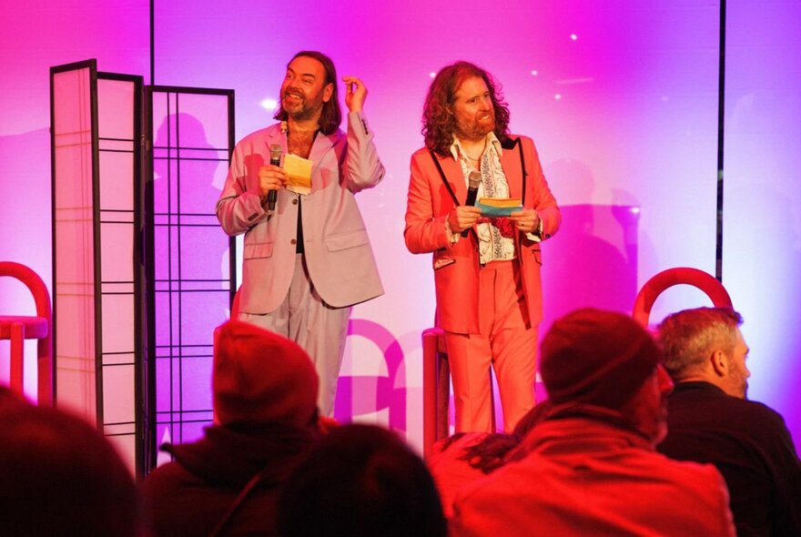 Presenters on stage holding cards and microphones against hot pink background.
