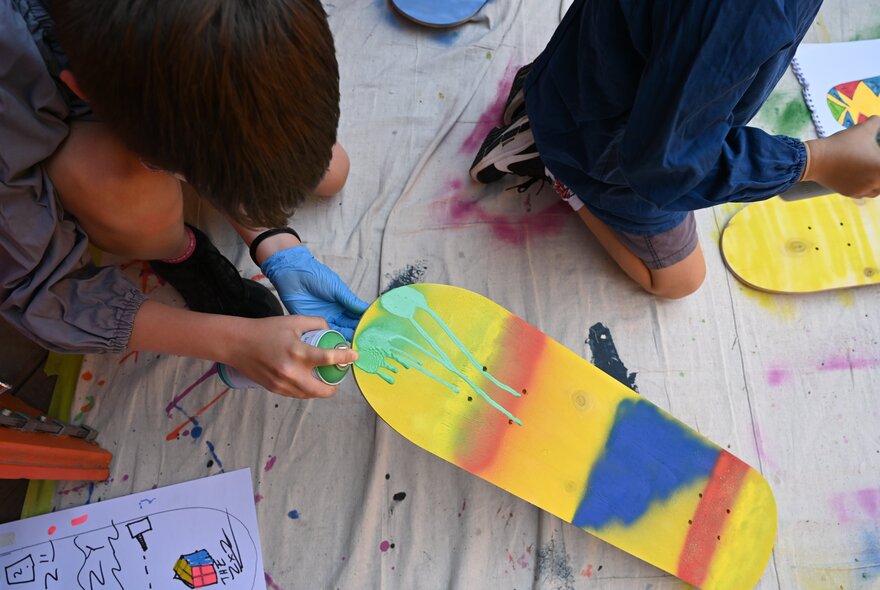 Children using spray cans and stencils to decorate skateboard decks in an art workshop.