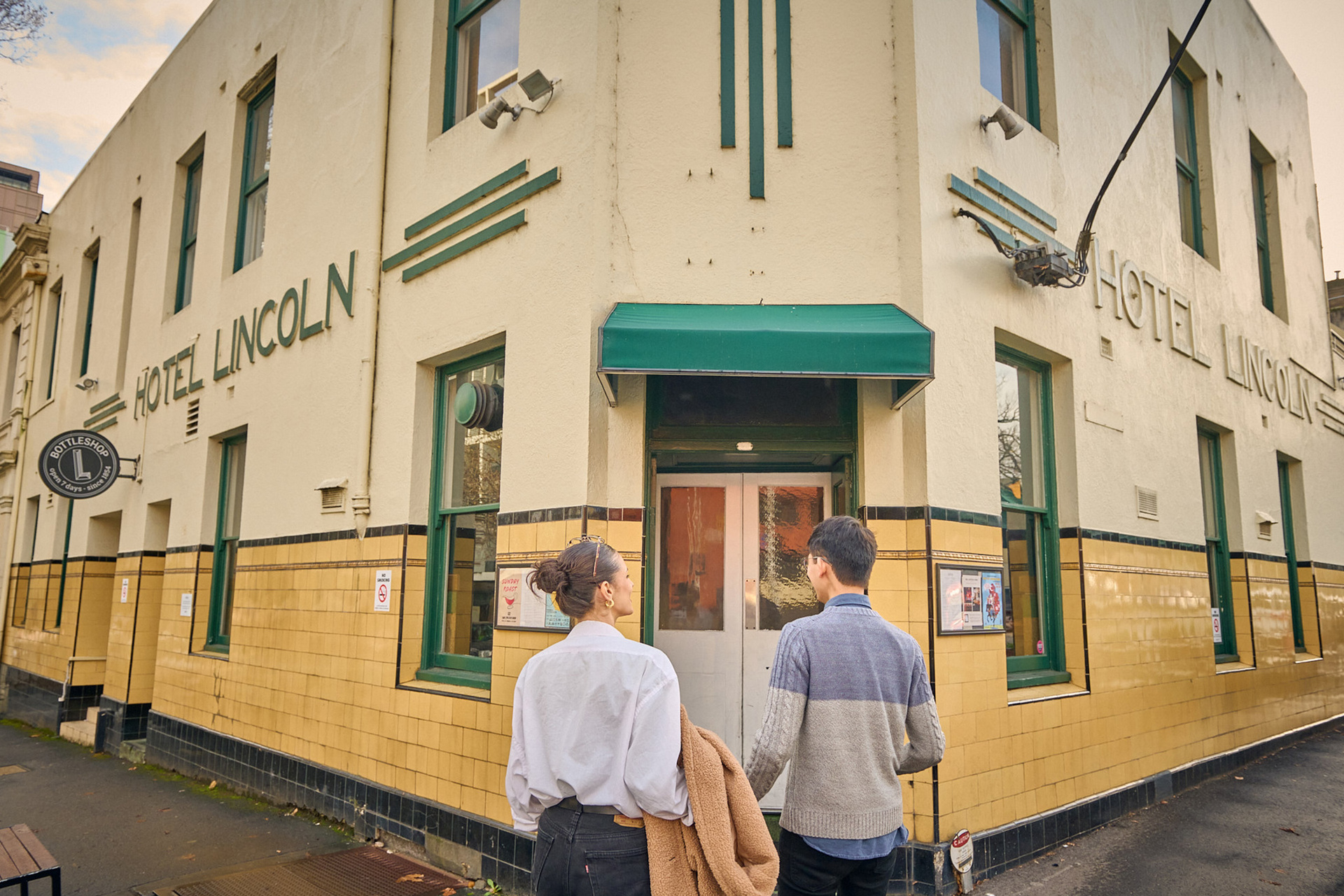 A couple are sitting walking into an art deco style corner pub there is a sign on the outside saying hotel lincoln