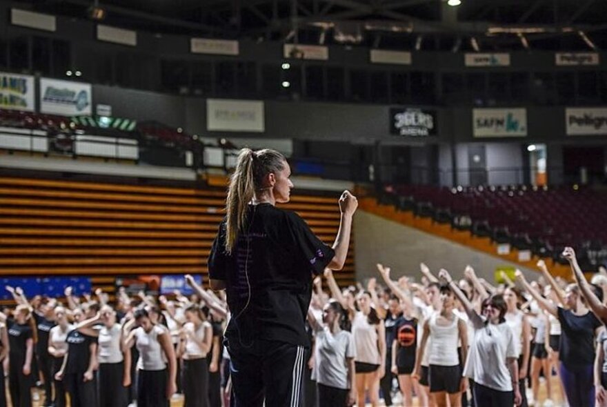 Choreographer Stephanie Lake teaching a large group of dancers inside a stadium.
