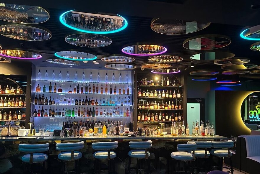 Large cocktail bar, with a row of high bar stools facing the bar counter, and a back wall of bottles of alcohol, and a ceiling with mirrors.