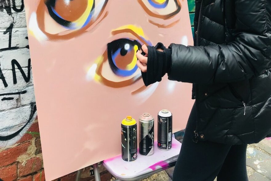 A person wearing dark clothes decorating stencil art with cans on a canvas in a laneway.