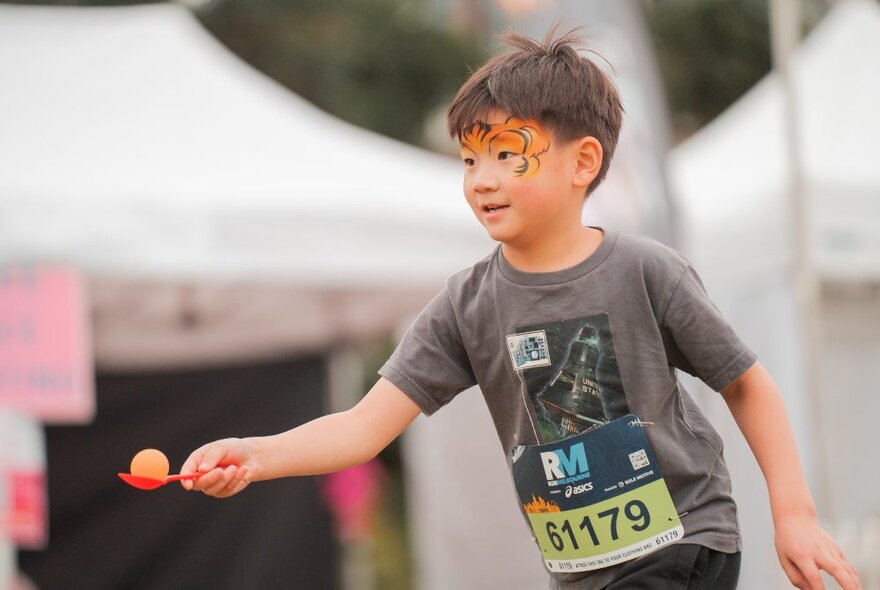 A small boy with face paint participating in an egg and spoon race with an event number on his chest. 