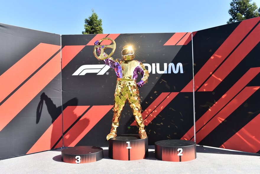 F1 driver in gold suit holding a gold steering wheel aloft.