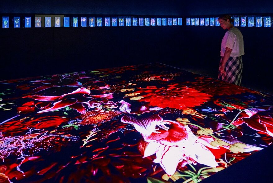 A woman is in a gallery looking at a neon-lit projection of flowers