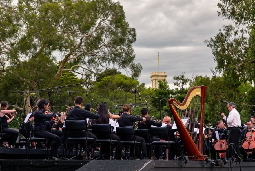 Looking at the back of an orchestra playing live on stage in the Royal Botanic Gardens Melbourne, with a view of the trees and the tower of Government House in the background.