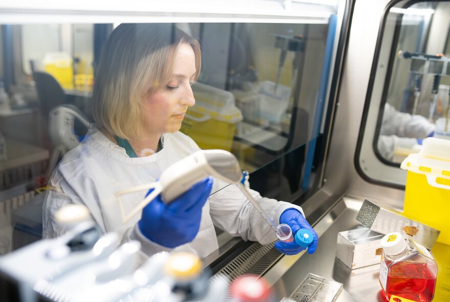 Researcher in white gown and blue gloves, working within a specialised biosafety cabinet in a laboratory setting.