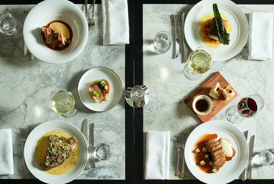 Overhead view of four plates of food arranged on a marble-topped dining table, with cutlery, cloth napkins and glasses of wine.