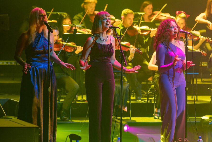 Three singers standing at microphones with seated violinists in the background.