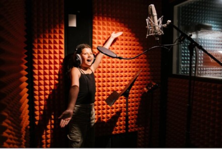 Child wearing headphones, singing with arms outstretched in front of a hanging microphone in a recording studio setup.