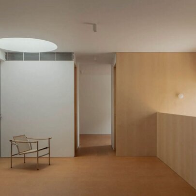 A stark room with blond timber on one side and a white wall on the other with a round ceiling light and single chair. 
