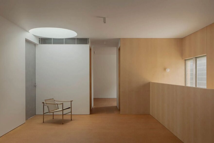 A stark room with blond timber on one side and a white wall on the other with a round ceiling light and single chair. 