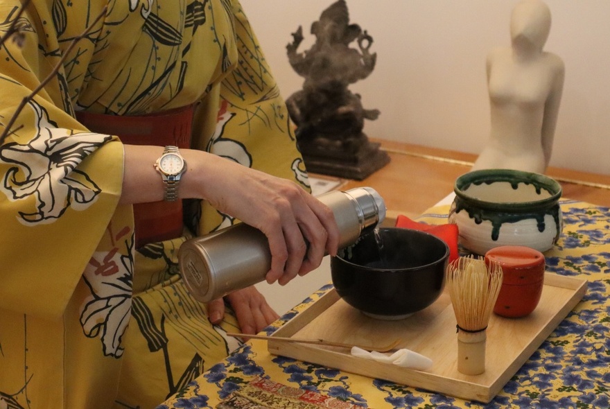 A Japanese woman wearing a dark yellow kimono, pouring tea from a flask into a small black bowl on a wooden tray, with a traditional tea whisk.