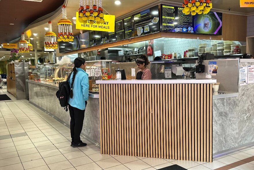 The bar in an Indian Restaurant, with a customer in blue talking to a worker behind the counter.