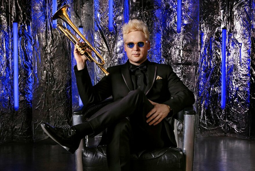 Musician Matt Von Roderick sitting cross-legged in a chair holding a trumpet up against a vivid blue and black background. 