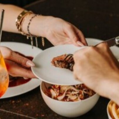 Hands reaching for shared dishes of food over a restaurant table, with iced drink in a wine glass with straw.