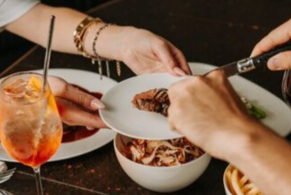 Hands reaching for shared dishes of food over a restaurant table, with iced drink in a wine glass with straw.