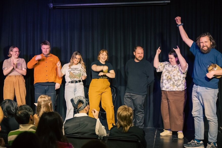 A cast of impro theatre performers on a stage in front of a seated audience.