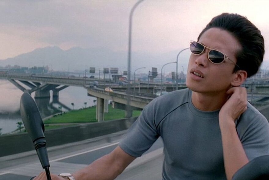 Film still of a man wearing a grey top and sunglasses, seated outside with a highway and a mountain in the background.