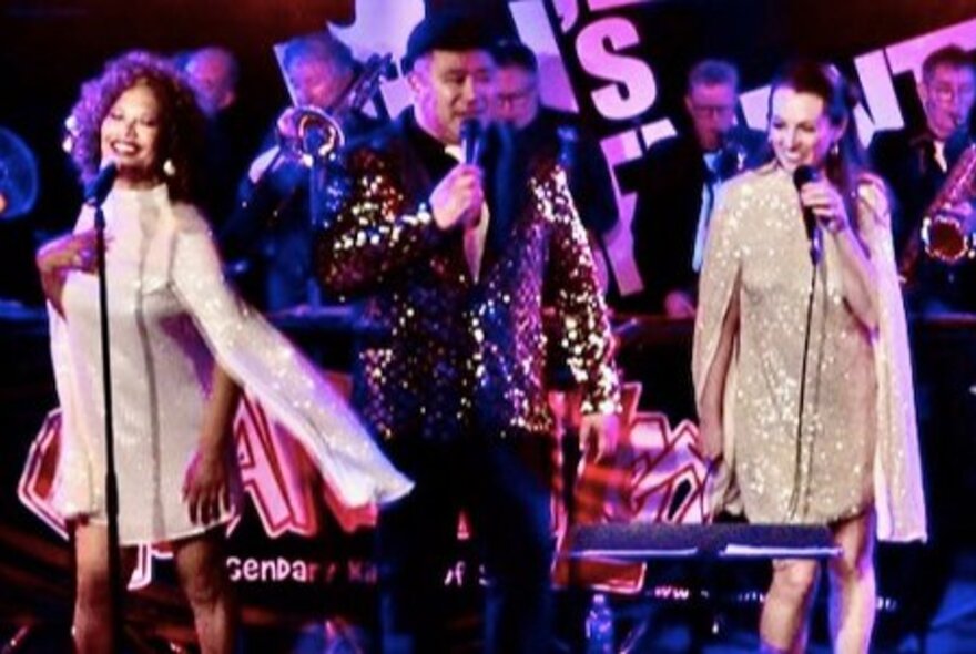 Three members of the band Grand Wazoo taking centre stage and performing, with a big band playing behind them.