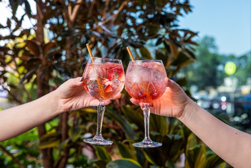 Two arms and hands holding large glasses of an icy drink with straws, clinking them together in a celebratory way.