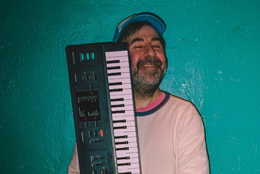 David O'Doherty, a smiling man with a short beard, holding up a digital keyboard in front of his chest, against a painted green wall.