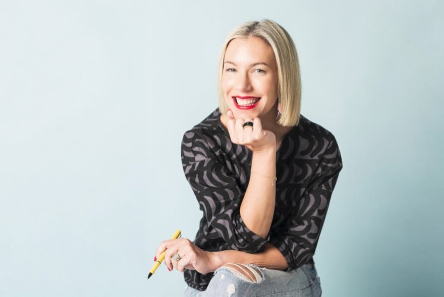 A woman seated with her legs cross and her chin leaning on her hand, smiling with a pen in her hand. 