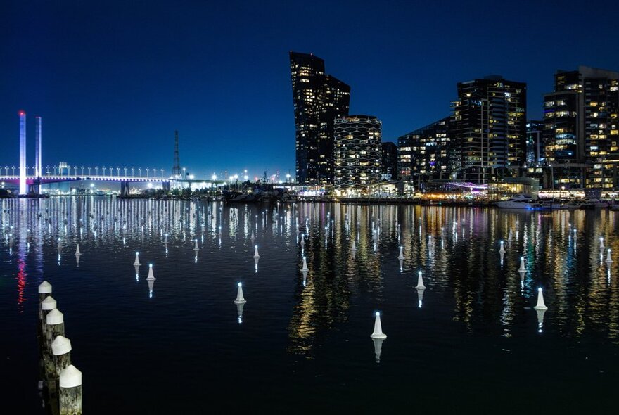 Night aerial view of the Melbourne Docklands area and city skyline, with the calm dark waters of Victoria Harbour and a public art installation of illuminated buoys in rows in the foreground. 