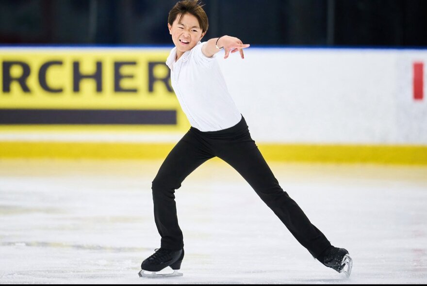 A young male figure skater in black pants and a white shirt, skating on ice.