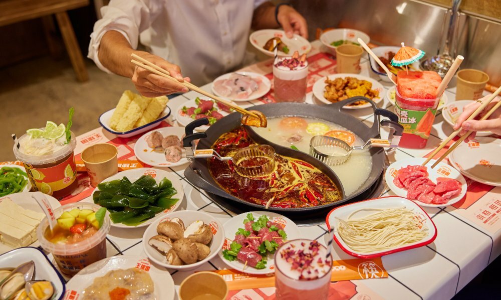 Two friends are eating Chinese hotpot there are many plates of meat, vegetables and seafood on the table.