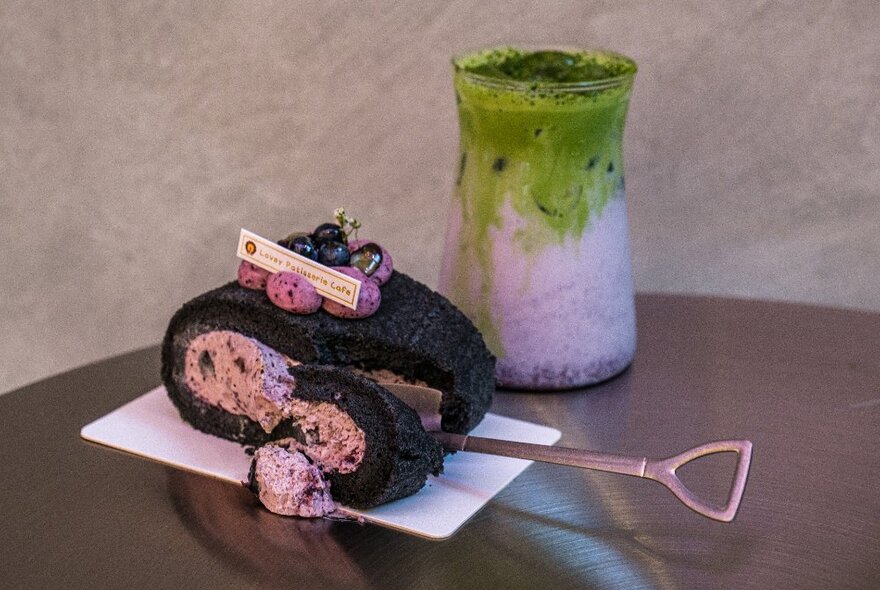 Blueberry elderflower swiss roll cake and an iced matcha drink on a stainless steel table.