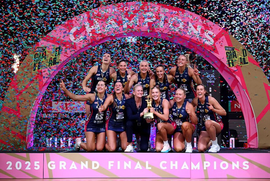 The Melbourne Vixens team celebrating their victory as the 2025 Suncorp Super Netball Champions, under a banner that reads CHAMPIONS, with tinsel confetti in the air around them.