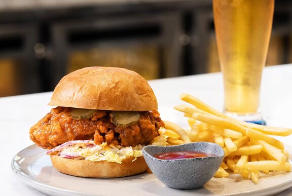 A chicken burger on a plate with fries and a small bowl of sauce, a schooner of icy cold beer behind the plate.