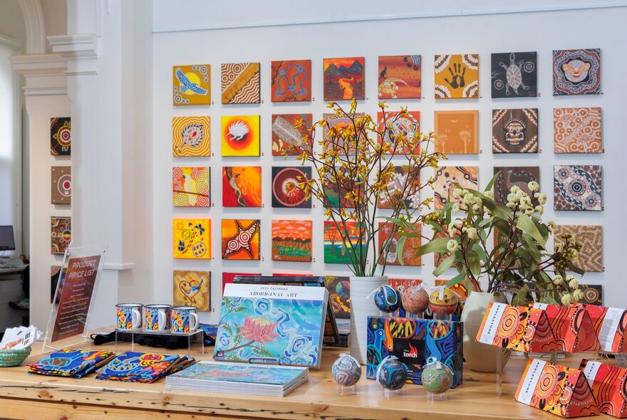 A gallery with paintings arranged on a wall and a table in the foreground with books, catalogues, postcards and a vase of flowers.