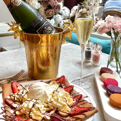A brunch table setting with a plate of strawberry chocolate crepes with ice cream, a platter of  macarons, a flute of champagne, and a bottle of champagne resting in a gold ice bucket on a marble tabletop.