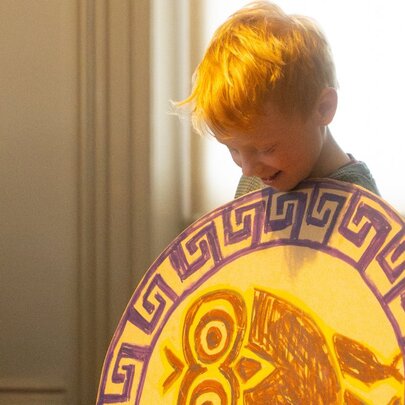 A child holding a hand-drawn Greek shield in front of his chest as he looks down at it.