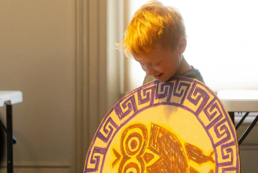 A child holding a hand-drawn Greek shield in front of his chest as he looks down at it.