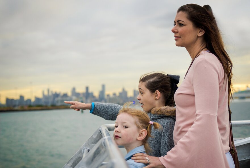 A mother with her two young children standing on the deck of a boat cruising the Yarra River, with one of the children smiling and pointing at something we can't see.