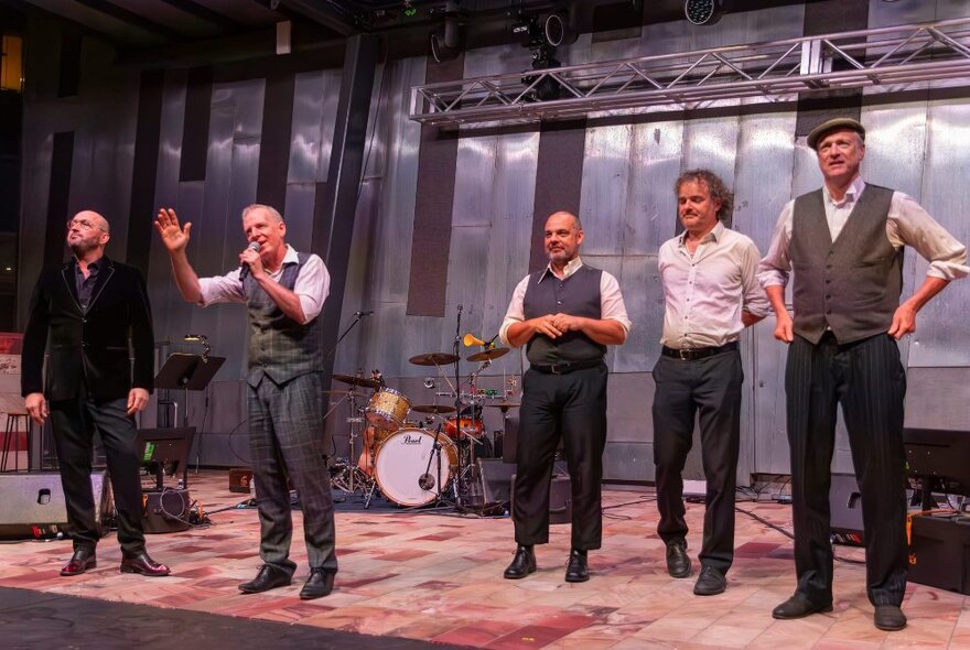 Five men on stage with a drumkit, wearing suits pants, shirts and vests, one with a microphone.